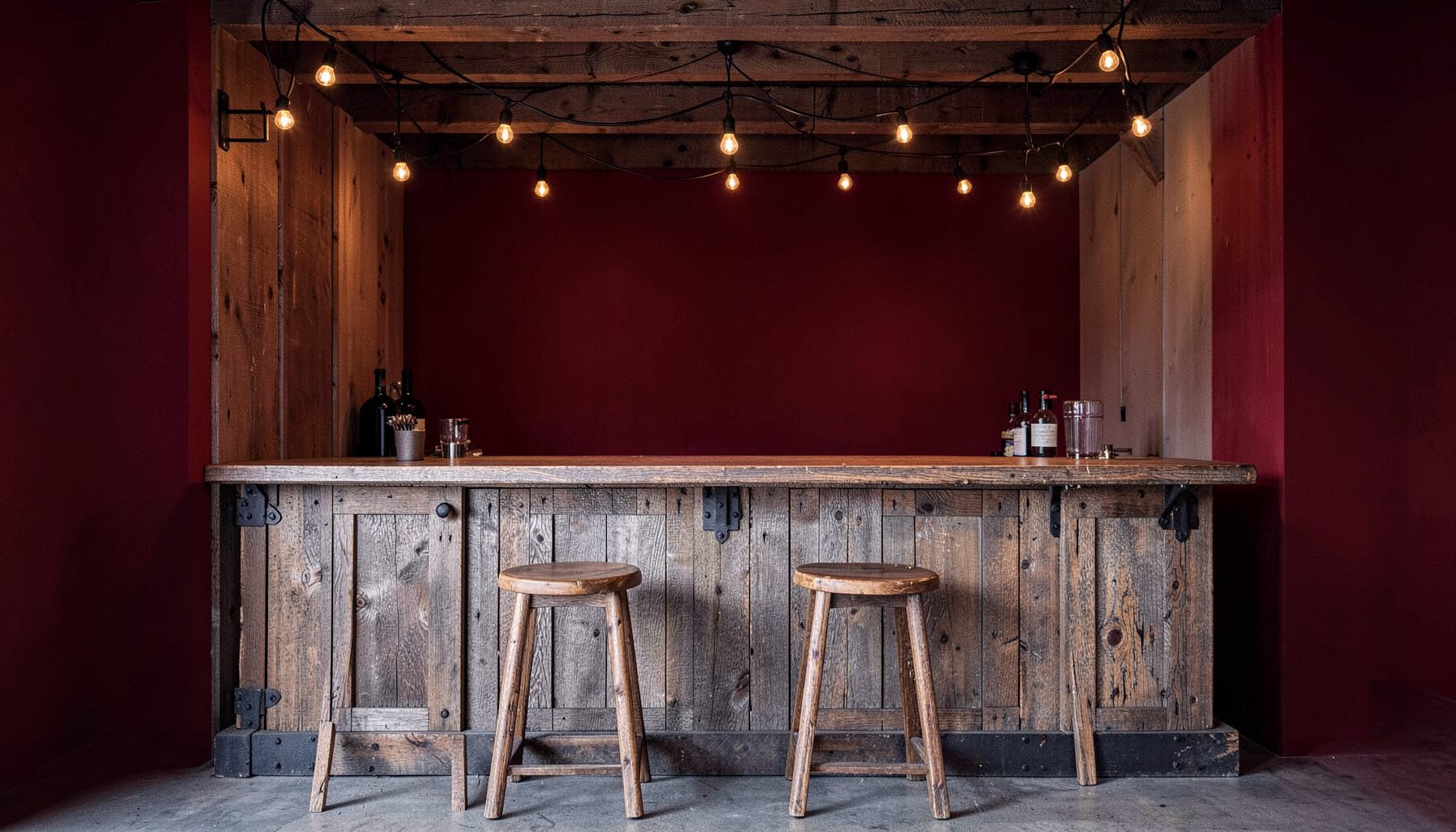 A weathered wooden rustic bar with iron accents, warm string lights above, and wooden stools in front.