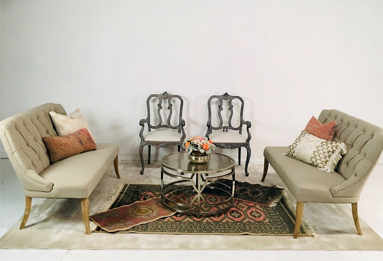 Two tufted beige settees facing each other with ornate dark wood chairs, a round glass coffee table, and decorative pillows in an elegant lounge setting with warm neutral tones