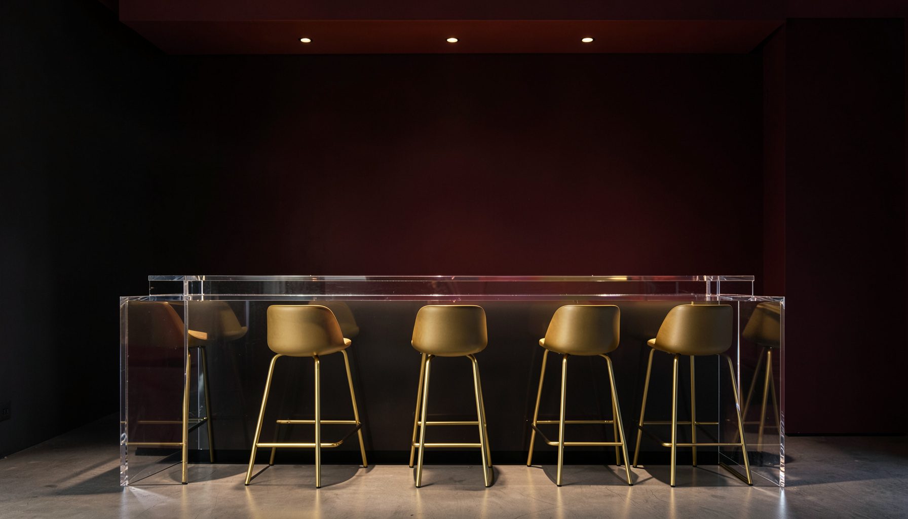 A sleek modern bar setup with a clear Lucite counter, gold bar stools, and warm ambient lighting against a dark wall.