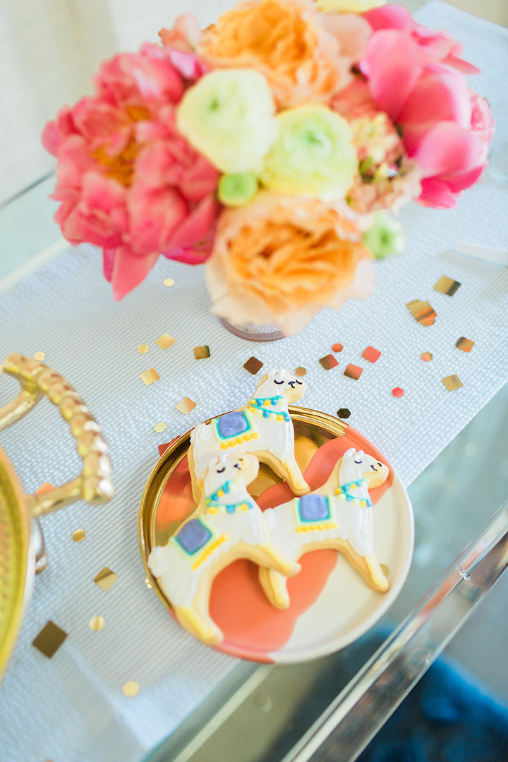 Close-up of decorated llama-shaped cookies on a gold plate beside a colorful floral arrangement, with gold confetti scattered around, warm and festive mood
