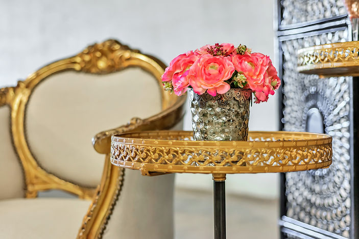 Ornate gold side table holding a metallic vase with pink and coral flowers beside an elegant upholstered chair
