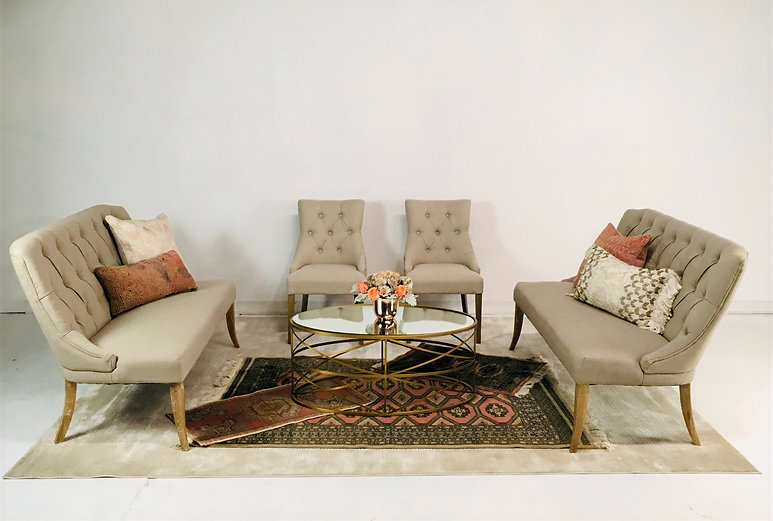 Two tufted beige settees with matching chairs, a round glass-top gold coffee table, and decorative pillows in a refined lounge