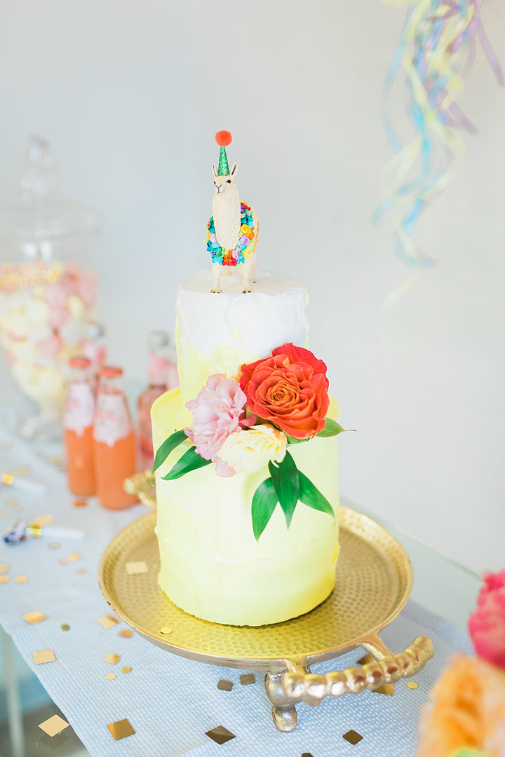 Pale yellow two-tier cake topped with a small llama figurine wearing a party hat, decorated with fresh red and pink flowers