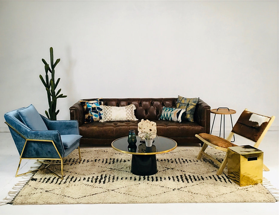 A styled living room vignette with a brown leather tufted sofa, blue velvet armchair, gold accents, patterned pillows, and a glass coffee table.